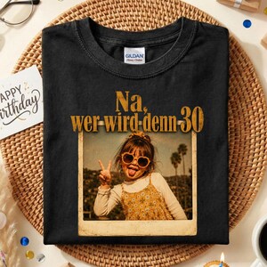 May include: A black t-shirt featuring a graphic print of a child wearing heart-shaped sunglasses and a floral dress. The text above the image reads, "Na, wer-wird-denn-30". The shirt has a Gildan brand label on the inside of the collar.