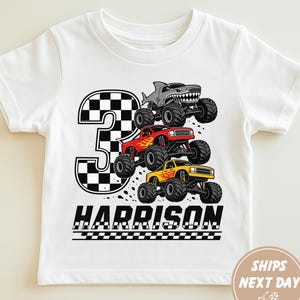 May include: A white short-sleeve t-shirt featuring a large black and white checkered number three, three monster trucks in gray, red, and yellow, and the name Harrison printed in black capital letters above a checkered racing flag pattern.
