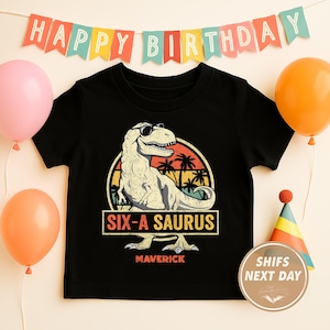 May include: Black t-shirt featuring a dinosaur wearing sunglasses with the text "SIX-A SAURUS" and "MAVERICK". The shirt is decorated with a colorful sunset design and is surrounded by balloons and a birthday banner.