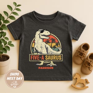 Op de afbeelding: Donkergrijs T-shirt met een dinosaurus met zonnebril, de tekst "FIVE-A SAURUS" en de naam "Harrison". Het ontwerp is geplaatst tegen een retro zonsondergang achtergrond. Inclusief een paar bruine schoenen en een houten schommelpaard.