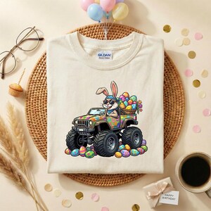 May include: Cream-colored t-shirt featuring a cartoon Easter bunny driving a monster truck filled with Easter eggs. The truck is decorated with colorful patterns. The bunny wears sunglasses and a bandana. Easter-themed apparel.