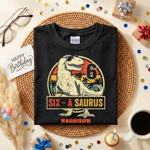 Pode incluir: Camiseta preta com um desenho de dinossauro usando óculos de sol, o número 6 e o texto "SIX-A SAURUS". O nome "Harrison" está impresso abaixo. O design é colocado em um fundo de pôr do sol. Tema de aniversário.