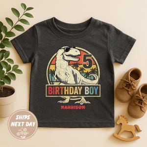 May include: Dark gray t-shirt featuring a dinosaur wearing sunglasses, with the number 5 and the text "BIRTHDAY BOY" in a retro design. The name "Harrison" is printed below. The shirt is next to a pair of brown shoes and a wooden rocking horse.