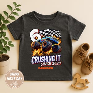 Puede incluir: Camiseta gris oscuro con un diseño de camión monstruo colorido, el texto "Crushing It Since 2020" y el nombre "Harrison". El diseño incluye una bandera a cuadros y llamas, celebrando un sexto cumpleaños.