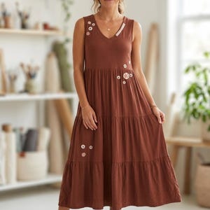 Peut inclure: Une robe longue à étages de couleur brun rouille, sans manches, avec une encolure en V et des boutons décoratifs dispersés de différentes tailles et nuances de blanc et de brun sur le corsage et la jupe, portée avec des sandales à lanières.