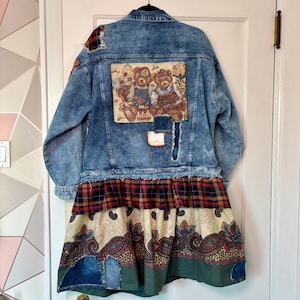 Może przedstawiać: Unikatowa kurtka dżinsowa z patchworkiem. Kurtka ma jasnoniebieskie pranie, postrzępione detale i dekoracyjną gobelin na plecach z misiami. Dół kurtki to warstwowa spódnica z wzorami w kratę i paisley.
