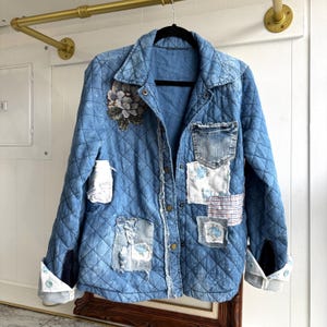 Op de afbeelding: Een lichtblauwe spijkerjas met een gewatteerd patroon en diverse stoffen patches. De jas heeft een bloemenapplicatie en knoopsluitingen. De manchetten zijn opgerold en tonen een wit en blauw gevoerde voering. De jas hangt aan een zwarte hanger.