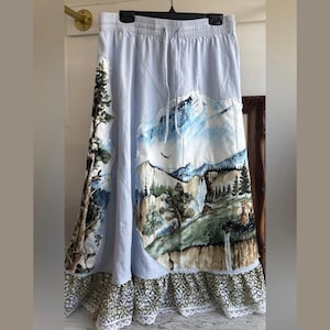 May include: A light blue skirt with an elastic drawstring waistband, featuring a large patch depicting a mountain landscape with trees and a waterfall. The hem is finished with a wide, patterned green and white fabric border and white lace trim.