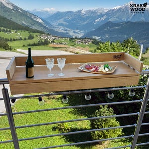 May include: A wooden balcony table with a bottle of wine, two wine glasses, and a plate of food. The table is mounted on a metal railing with a scenic mountain view in the background. The table has the text "KRAS WOOD DESIGN".