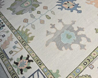 May include: A cream-coloured area rug with a floral pattern in shades of blue, green, pink, and grey. The rug features a border with repeating floral and geometric designs. The overall design is a traditional, yet modern, style.
