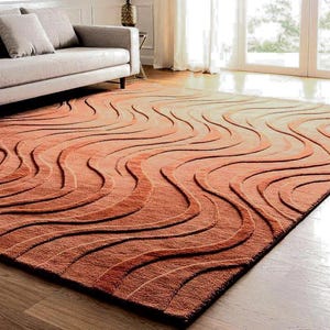 May include: A large, rectangular rug with a wave-like pattern in shades of orange and brown. The rug is placed on a wooden floor, and a sofa is partially visible in the background. The rug's design adds a modern touch to the room.