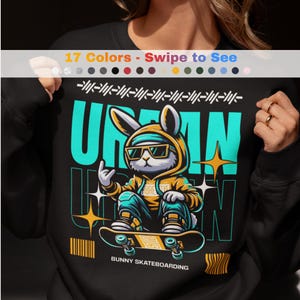 Puede incluir: Sudadera negra con un conejo de dibujos animados con gafas de sol y una sudadera con capucha, montando en monopatín. El diseño incluye la palabra "URBAN" en color turquesa, con estrellas y líneas. El texto "BUNNY SKATEBOARDING" está en la parte inferior.