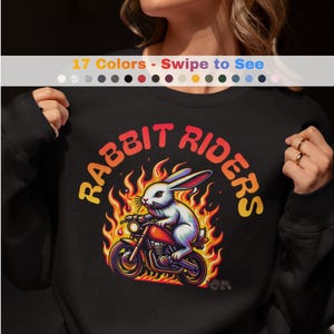 Puede incluir: Sudadera negra con un gráfico de un conejo en una motocicleta rodeado de llamas. El texto "RABBIT RIDERS" está encima de la imagen en naranja y amarillo. El diseño está centrado en la parte delantera de la sudadera.
