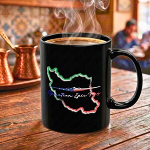 Op de afbeelding: Een zwarte koffiemok met een handvat, gevuld met koffie en stoom die van bovenaf opstijgt. De mok heeft een kaartomtrek met groene, rode en blauwe accenten en de tekst "Operation Epic Day".