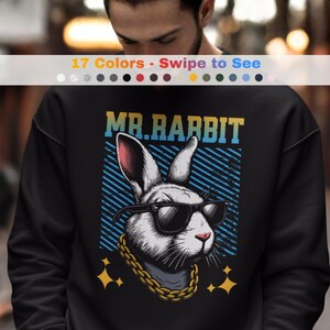 Puede incluir: Sudadera negra con un gráfico de un conejo con gafas de sol y una cadena de oro. El texto "MR. RABBIT" está encima del conejo. La sudadera tiene cuello redondo y mangas largas. La imagen también incluye el texto "17 Colors - Swipe to See".