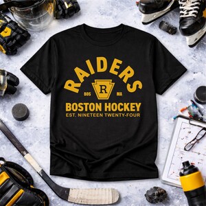 May include: Black t-shirt with gold text reading "RAIDERS" and "BOSTON HOCKEY". The graphic includes a keystone shape with the letter "R" inside. The text "EST. NINETEEN TWENTY-FOUR" is below the graphic. Hockey equipment, including skates and a stick, surrounds the shirt.