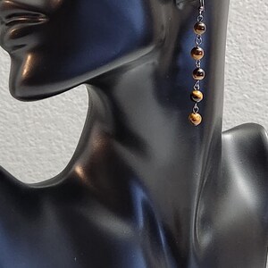 May include: A pair of dangle earrings featuring five round tiger's eye beads. The beads are strung on a delicate silver-tone chain. The earrings are displayed on a black mannequin head against a neutral background.