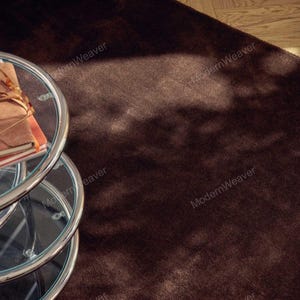 May include: A dark brown, plush rug with a soft texture. A glass-topped, three-tiered side table with a chrome frame is in the foreground, with a stack of books on the top tier. The rug covers most of the frame.