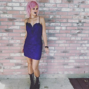 Puede incluir: Una mujer con el pelo rosa y labios oscuros lleva un vestido corto de encaje morado y botas negras con tachuelas. Está de pie frente a una pared de ladrillo.