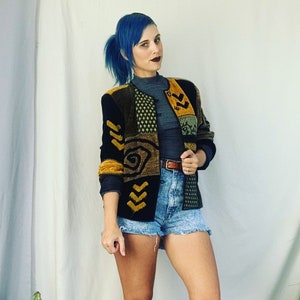 Könnte beinhalten: Eine Frau mit blauen Haaren trägt eine schwarze, grüne und gelbe gemusterte Strickjacke mit einem braunen Gürtel und Jeans-Shorts.