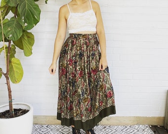 May include: A woman wearing a white lace crop top and a long, flowing floral skirt. The skirt has a green, brown, and red floral pattern and a dark green trim.