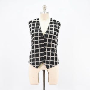 May include: A sleeveless vest featuring a black and white grid pattern. The garment is covered in small, reflective beads that form a checkerboard design. It has a deep V-neckline and a front button closure with three dark, circular buttons.
