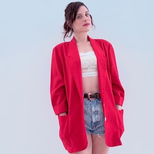 Könnte beinhalten: Ein leuchtend roter Blazer mit großen Fronttaschen und einem einzelnen Knopfverschluss. Der Blazer wird über einem weißen Spitzentop und Jeansshorts getragen. Ein brauner Gürtel mit silberner Schnalle betont die Taille.