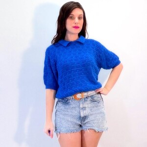 May include: A bright blue short-sleeved sweater with a textured pattern. The sweater has a polo collar and is paired with acid wash denim shorts and a decorative belt. The outfit is set against a plain white background.