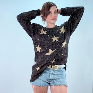 Op de afbeelding: Een zwart gebreide trui met lange mouwen en een herhalend patroon van gouden sterren en kleine zilveren lovertjes, gecombineerd met lichtblauwe acid-wash denim shorts met een gerafelde zoom en een witte riem met een gouden gesp.