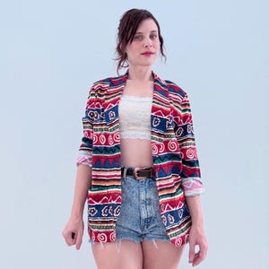 May include: A colorful, patterned blazer with rolled-up sleeves. The jacket features a mix of red, blue, green, and white geometric designs. The model is wearing denim shorts and a white top. A black belt is visible.