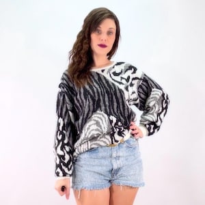 May include: A vintage sweater with a black, white, and gray animal print design. The sweater has a crew neck and long sleeves with white cuffs. The model is wearing light blue denim shorts and a gold belt.