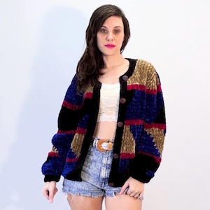 Op de afbeelding: Een veelkleurig vest met knopen en een textuurrijke, zachte stof. De jas heeft een colorblock-ontwerp met zwarte, blauwe, bruine en rode strepen. Het heeft een ronde halslijn en donkerbruine knopen. De mouwen zijn lang.