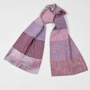 Könnte beinhalten: Rechteckiger Schal in Lila-, Lavendel- und Mauve-Tönen. Der Schal weist eine Vielzahl von Mustern auf, darunter Streifen, strukturierte Abschnitte und ein Design mit wirbelnden grauen Linien. Der Schal ist aus einem weichen, fließenden Stoff gefertigt.
