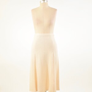 May include: A beige, knee-length skirt displayed on a mannequin. The skirt has a fitted waist and a slight A-line shape. Vertical seams and decorative cutouts add detail to the front panels. The fabric appears to be a soft knit.