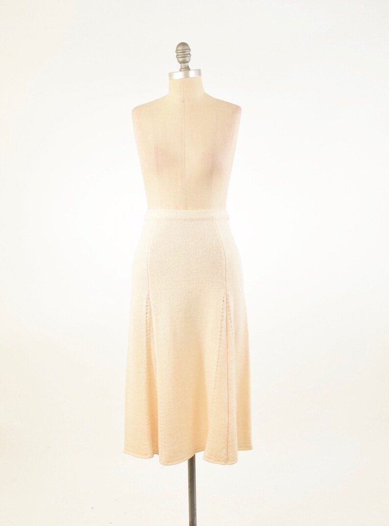 May include: A beige, knee-length skirt displayed on a mannequin. The skirt has a fitted waist and a slight A-line shape. Vertical seams and decorative cutouts add detail to the front panels. The fabric appears to be a soft knit.