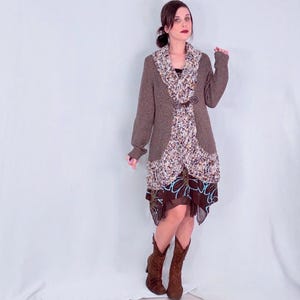 Pode incluir: Um cardigã de malha castanho de manga comprida com fecho de alavanca sobre um vestido em camadas. O vestido apresenta um corpete com estampado floral e uma saia castanha escura com um padrão de flores azul-claro e botas de camurça castanhas.