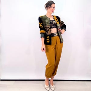May include: A long-sleeve jacket featuring a black, gold, and olive green patchwork pattern with chevron details, worn over a black lace crop top, paired with high-waisted mustard yellow trousers, a black belt with a gold buckle, and white shoes.