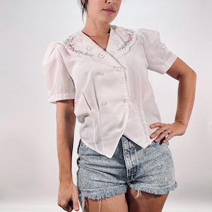 Op de afbeelding: Witte blouse met korte mouwen en een dubbelknoops ontwerp. De blouse heeft een decoratieve kraag met bloemenborduurwerk en een geschulpte rand. De blouse wordt gecombineerd met een spijkerbroek met zuurwassing.