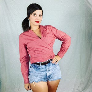 May include: A woman wearing a pink wrap shirt with long sleeves and a brown belt. She is also wearing denim shorts.