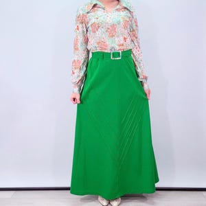 Puede incluir: Una falda larga verde vintage con cinturón a juego y una blusa con estampado floral. La falda tiene cintura alta y una silueta larga y fluida. La blusa tiene cuello y mangas largas. El conjunto es de estilo retro.