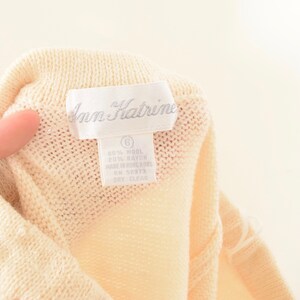 May include: Close-up of a cream-colored knit garment with a label that reads "Ann Kathrine." The label also indicates the garment is size 6 and made of wool and rayon. The garment is dry clean only.