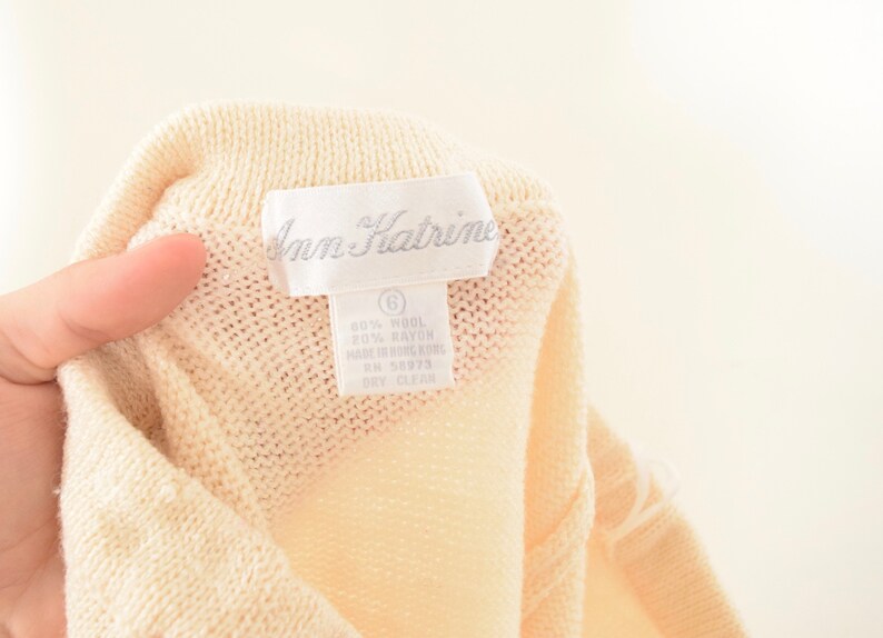 May include: Close-up of a cream-colored knit garment with a label that reads "Ann Kathrine." The label also indicates the garment is size 6 and made of wool and rayon. The garment is dry clean only.