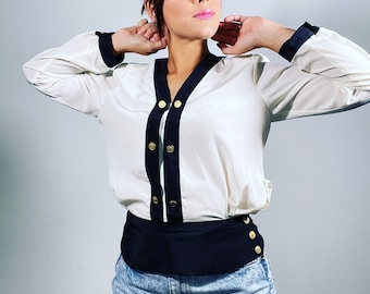 May include: A white button-up shirt with black trim and gold buttons. The shirt is worn with a black belt and denim shorts.