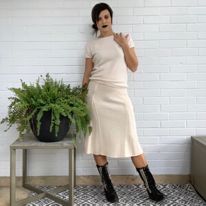 May include: A cream-colored knit skirt and short-sleeved top set. The skirt has a flared hem and vertical stitch detailing. The top has a pocket on the chest. The person is wearing black boots with silver studs. A potted fern sits on a side table.