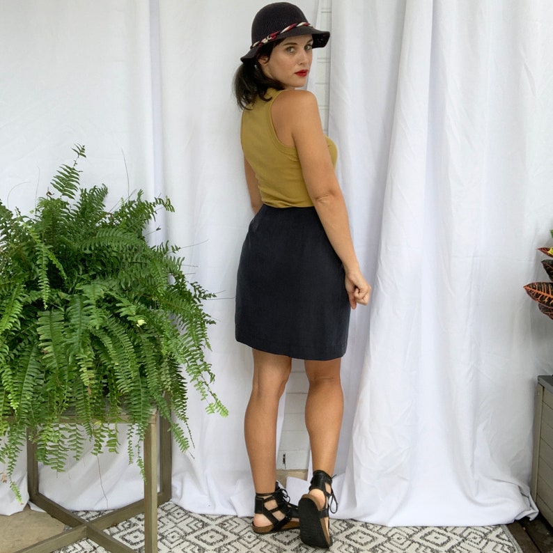 May include: A woman wearing a mustard yellow sleeveless top, a dark blue mini skirt, and black sandals. She is wearing a black and white patterned hat and is standing in front of a white curtain.