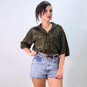 Könnte beinhalten: Ein kurzärmeliges, olivgrünes Hemd mit Knopfleiste, leichtem Glanz und einer Brusttasche, kombiniert mit einer hoch taillierten Acid-Wash-Jeansshorts mit ausgefranstem Saum und einem dünnen braunen Ledergürtel mit goldfarbener Schnalle.