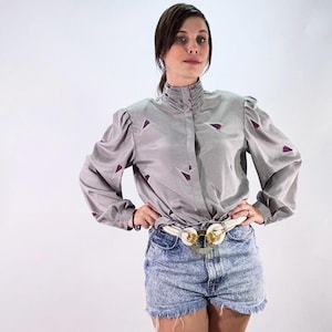 May include: A gray long-sleeve blouse with a high collar and burgundy triangle accents. The blouse is paired with acid wash denim shorts and a white belt with a gold buckle. The outfit is set against a white background.
