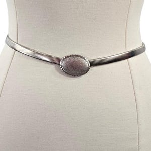 Könnte beinhalten: Ein silberfarbener Metallgürtel mit einer ovalen Schnalle. Der Gürtel besteht aus flexiblem, geripptem Material und wird auf einer Schaufensterpuppe präsentiert. Die Schnalle hat eine strukturierte Oberfläche.