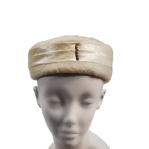 Puede incluir: Un sombrero de pastillero blanco con una banda de satén y una fila de diamantes de imitación.