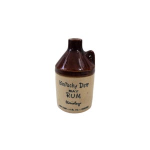 May include: A small, brown and tan ceramic jug with a handle. The jug is labeled "Kentucky Dew Bay Rum" in black text. The jug is 4 fl. oz.